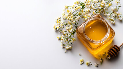 Overhead shot of golden honey in a glass jar with a honey dipper and delicate white flowers on a clean white background with copy space