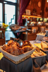 Hotel breakfast buffet with basket of croissants, assorted pastries, cakes, and desserts. Elegant self-service table with tongs, tablecloths, and warm lighting in a cozy dining area