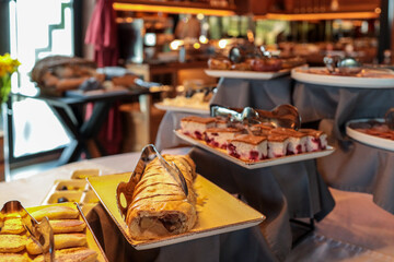 Hotel breakfast buffet with apple strudel, sponge cake with berries, cookies, and various desserts on plates with tongs. Elegant display of fresh pastries in cozy restaurant