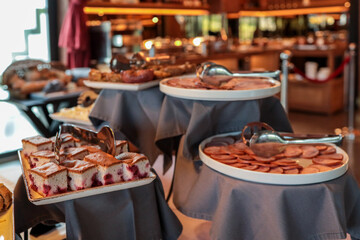 Buffet table with sliced salami, ham, sponge cake with berries, and other breakfast foods. Plates...