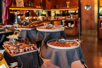Buffet table with sliced salami, ham, sponge cake with berries, and other breakfast foods. Plates with tongs ready for self-service in a hotel restaurant with warm lighting