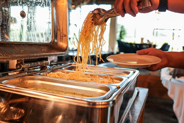 Close-up of a person serving hot spaghetti from a buffet chafing dish onto a plate. Warm lighting, steam on metal lid, fresh pasta ready for meal at restaurant or event catering