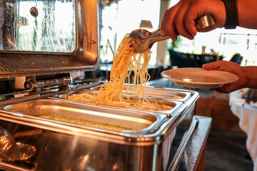 Close-up of a person serving hot spaghetti from a buffet chafing dish onto a plate. Warm lighting,...
