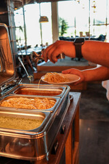 Close-up of a person serving hot spaghetti from a buffet chafing dish onto a plate. Warm lighting, steam on metal lid, fresh pasta ready for meal at restaurant or event catering