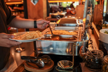 Buffet scene with person serving spaghetti and sauce onto plate. Stainless chafing dishes, fresh...