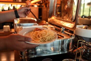 Close-up of person serving spaghetti with tomato sauce at buffet. Warm restaurant lighting,...