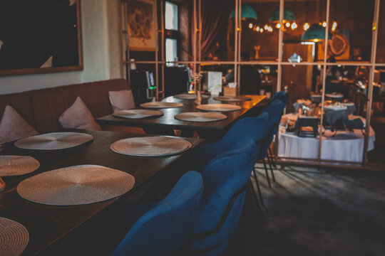 Cozy modern restaurant interior with dark wooden tables, blue velvet chairs, placemats, and soft lighting. Elegant dining space with a warm atmosphere and buffet area in the background