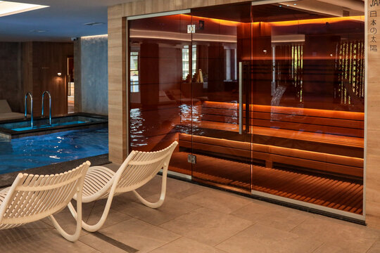 Modern spa area with illuminated sauna behind glass doors, lounge chairs and small indoor pool. Relaxing luxury wellness interior with wood, glass and stone design