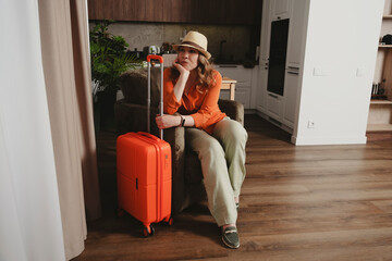 Woman awaits departure for summer vacation while sitting beside her orange suitcase in a cozy...