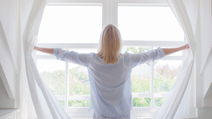 Rear View Of Woman Wearing Pyjamas Opening Curtains In Morning And Looking Out Of Window