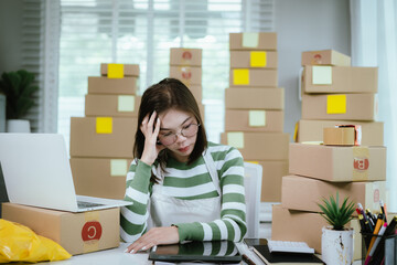 A stressed woman in a home office surrounded by shipping boxes uses a tablet, managing an online business with a laptop, calculator, and packing materials.