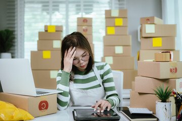 A stressed woman in a home office surrounded by shipping boxes uses a tablet, managing an online business with a laptop, calculator, and packing materials.