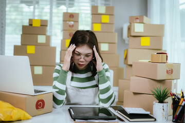 A stressed woman in a home office surrounded by shipping boxes uses a tablet, managing an online business with a laptop, calculator, and packing materials.