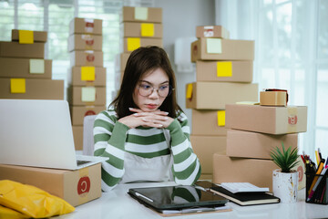 A stressed woman in a home office surrounded by shipping boxes uses a tablet, managing an online business with a laptop, calculator, and packing materials.