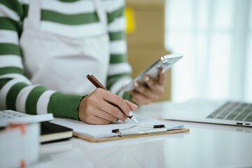 An Asian female entrepreneur works on her laptop to manage the logistics of her e-commerce business, organizing parcels, processing orders, and managing online sales and deliveries from her home offic