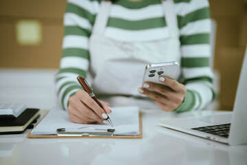 An Asian female entrepreneur works on her laptop to manage the logistics of her e-commerce business, organizing parcels, processing orders, and managing online sales and deliveries from her home offic