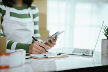 An Asian female entrepreneur works on her laptop to manage the logistics of her e-commerce business, organizing parcels, processing orders, and managing online sales and deliveries from her home offic