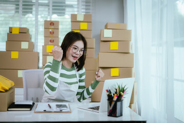 A young Asian woman entrepreneur in a striped shirt and apron works from her home office, managing e-commerce inventory, packaging orders, and organizing shipping for online customers.