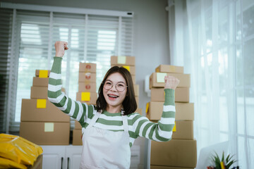 A young Asian woman entrepreneur in a striped shirt and apron works from her home office, managing...