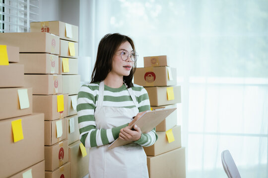 A young Asian woman entrepreneur in a striped shirt and apron works from her home office, managing e-commerce inventory, packaging orders, and organizing shipping for online customers.
