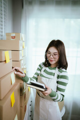 A young Asian woman entrepreneur in a striped shirt and apron works from her home office, managing e-commerce inventory, packaging orders, and organizing shipping for online customers.
