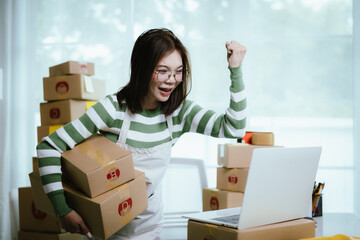 A young woman entrepreneur manages her online business from home, surrounded by parcel boxes, using...