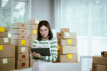 A young woman entrepreneur manages her online business from home, surrounded by parcel boxes, using...