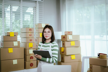 A young woman entrepreneur manages her online business from home, surrounded by parcel boxes, using...