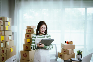 A young woman entrepreneur manages her online business from home, surrounded by parcel boxes, using a laptop, calculator, and notepad to handle shipping and inventory.