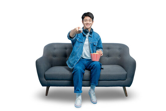 PNG,A young Asian man sits on a sofa with popcorn and points a TV remote at the camera, ready to start a movie.
