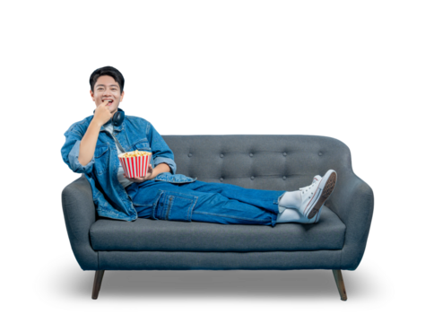PNG,An amused young man laughs and eats popcorn while lounging on a sofa, enjoying a comedy movie at home.
