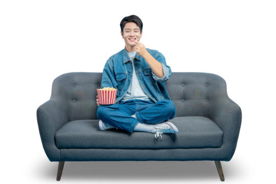 PNG,An amused young man smiles as he eats popcorn from a striped bucket while enjoying a show on his couch.
