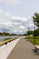 Naklejka premium Riverside pedestrian promenade with benches and streetlights along paved walkway under cloudy sky. 
