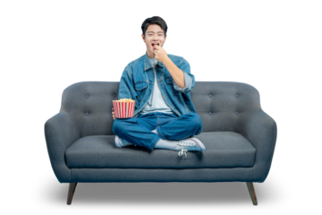 PNG,An amused young man smiles as he eats popcorn from a striped bucket while enjoying a show on his couch.
