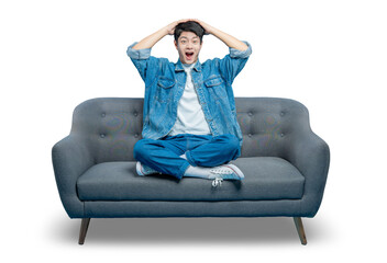 An amazed young man sits on a sofa with his hands on his head and mouth open in shock or...
