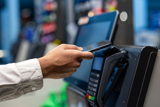 A person uses their smartphone to make a contactless payment at a point-of-sale terminal in a retail setting. The focus is on the hand and the device.