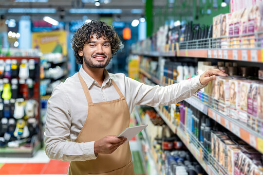 A smiling worker in a supermarket stands by shelves, checking inventory with a tablet while wearing an apron. - Powered by Adobe
