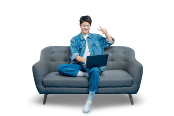 A happy young man makes a peace sign while using his laptop on a sofa, possibly during a video call or a study break.PNG