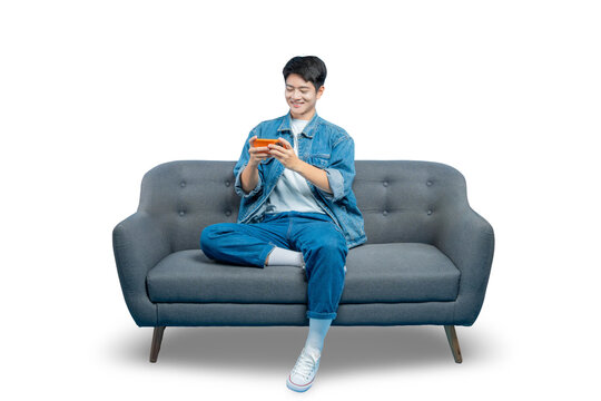 A happy young man in a denim jacket plays a game on his smartphone while relaxing on a sofa.PNG