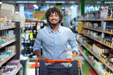 A smiling man pushes a shopping cart down an aisle in a brightly lit grocery store, focused on purchasing.