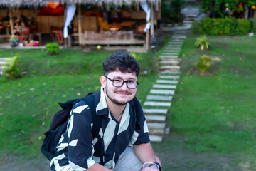 Traveler with backpack. Young man wearing stylish patterned shirt is sitting outdoors, surrounded...