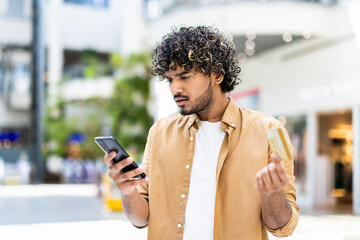 A man looking concerned while holding a credit card and smartphone in a shopping mall setting.