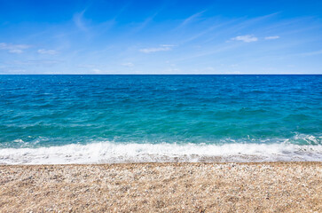 Pebble beach extends along calm turquoise sea beneath bright blue sky. Sea meets shore at clear...