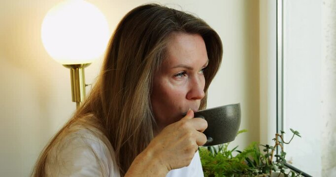 Mid aged woman drinks delicious latte from black porcelain cup in comfortable cafe. Lady guest takes sip of hot cappuccino from mug in coffeeshop
