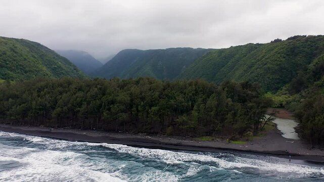 A winding river meets the crashing ocean waves along a dark sand shoreline bordered by dense forest, with steep green valley slopes rising into misty clouds in this dramatic wild landscape.