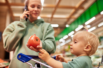 Happy young mother with her cute daughter in shopping trolley walking around supermarket. Beautiful...
