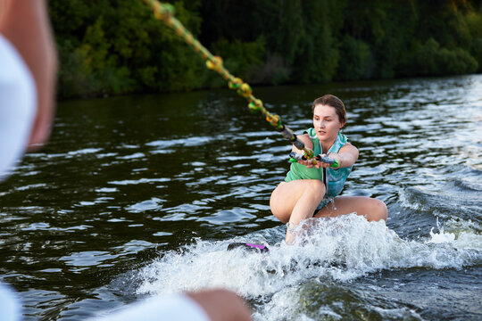 Young beautiful sporty girl in a green life jacket rides a wakeboard on a river or lake near city. Happy sportswoman glides on the wave. Active lifestyle, healthy hobby.
