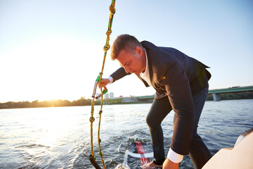 Young man in classic suit rides a wakeboard on the river or lake near city. Clerk escaped from a stuffy office to take up his favorite active sport. Best summer leisure after routine work. Sun flare.