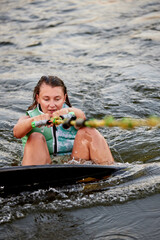 Young beautiful sporty girl in a green life jacket sitting in the water on a wakeboard with a...