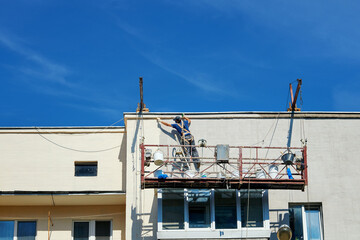 Unrecognizable construction worker on suspended scaffolding renovating facade of an apartment building, exterior maintenance for urban infrastructure improvement. Man restoring and painting facade